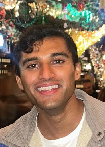 A young man with short dark hair and a bright smile, wearing a dark shirt, posing indoors near a window.