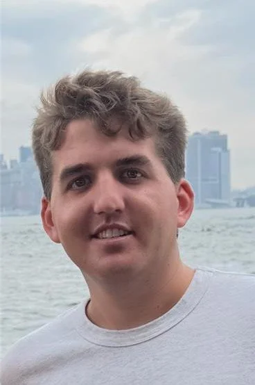 Young man with brown curly hair smiling outdoors with water and city skyline in the background.