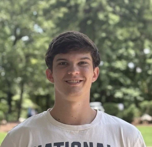 Young man smiling outdoors with trees in the background.