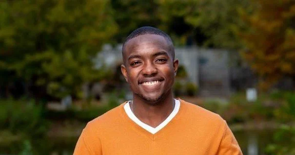 Smiling young man outdoors wearing an orange shirt with a white collar, with trees and water in the background.