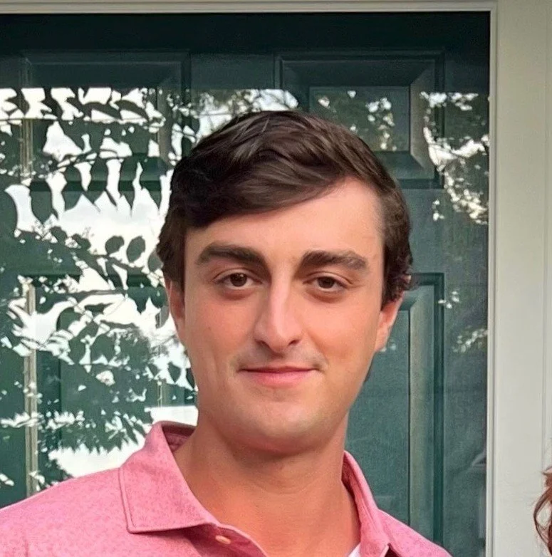 A young man with brown hair wearing a pink collared shirt, standing outside in front of a green door and window with reflection of trees.