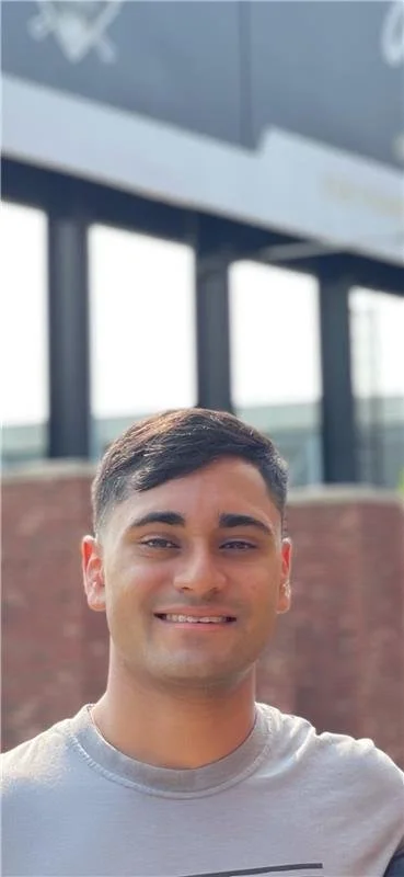 Young man smiling outdoors near a modern building with large windows and white signage in the background.
