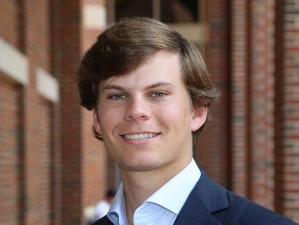 A young man with light brown hair, wearing a dark blazer and a light blue shirt, smiling outdoors with a brick building in the background.