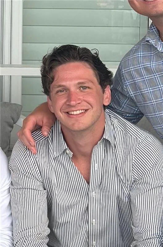 A young man with dark, curly hair smiling, wearing a black-and-white striped button-up shirt.
