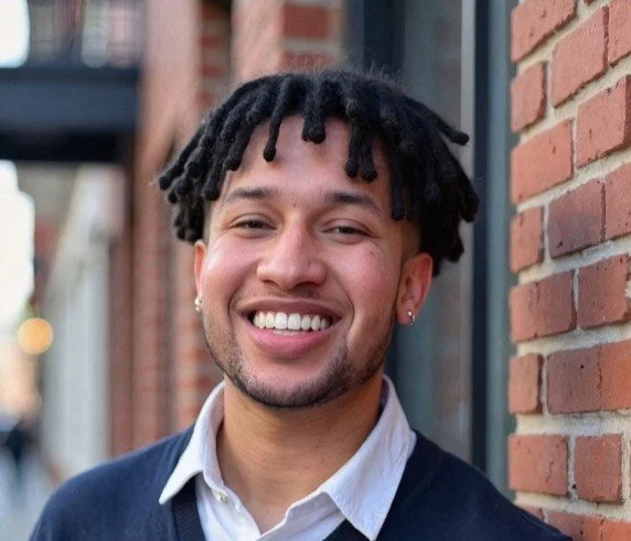 Young man with dreadlocks smiling outside in front of a brick wall.