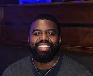 A smiling man with a beard and short hair, wearing a dark shirt, sitting in front of a wooden background.