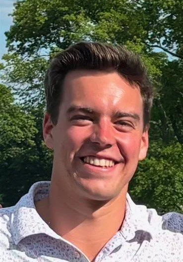 Smiling young man outdoors with green trees in the background.