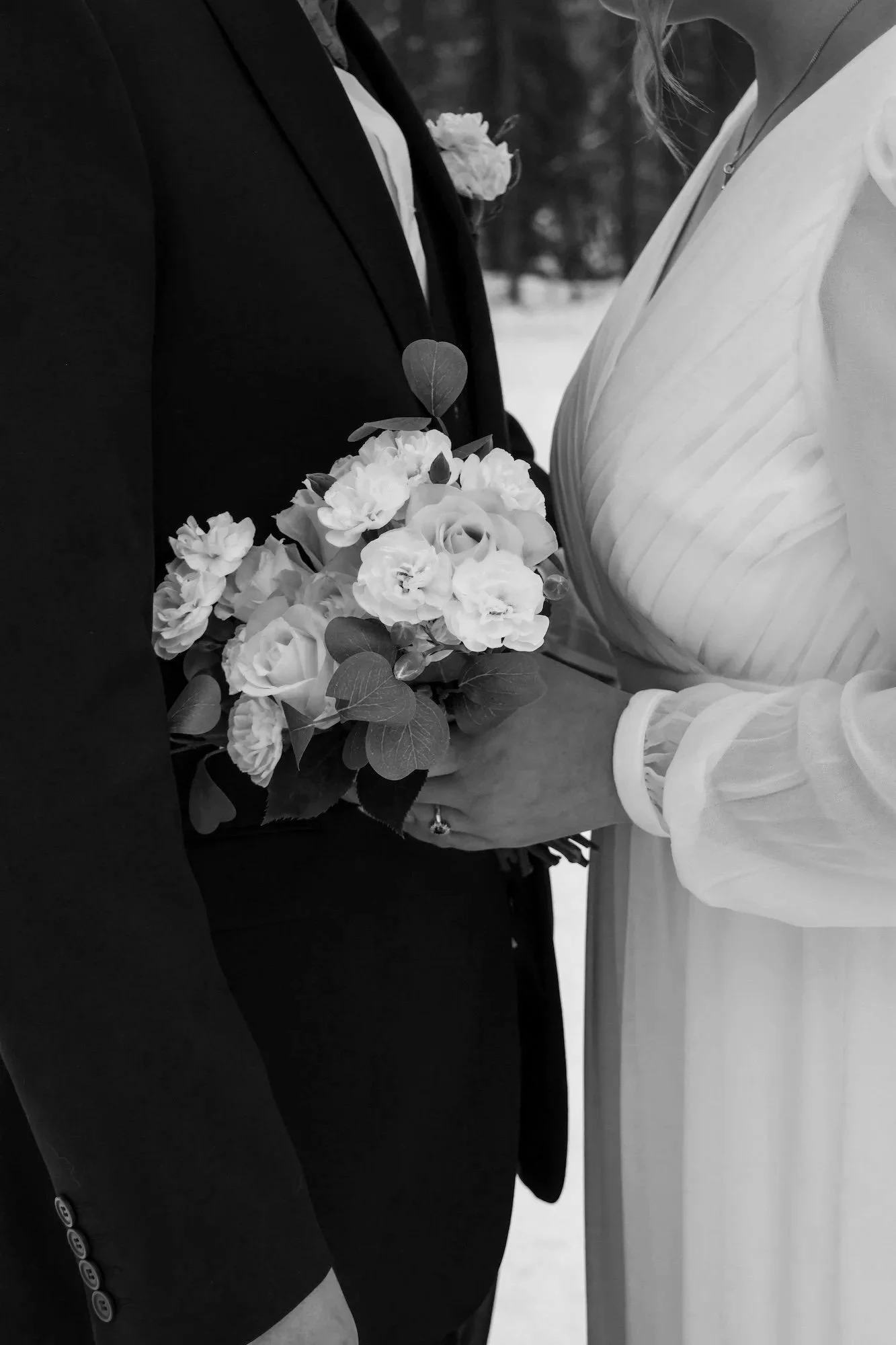 Hääkuvassa valkoiset kukat morsiuskimpussa hääparin keskellä, joka on pukeutunut pukuun ja hääpukuun, kuvattuna läheltä mustavalkoisena.