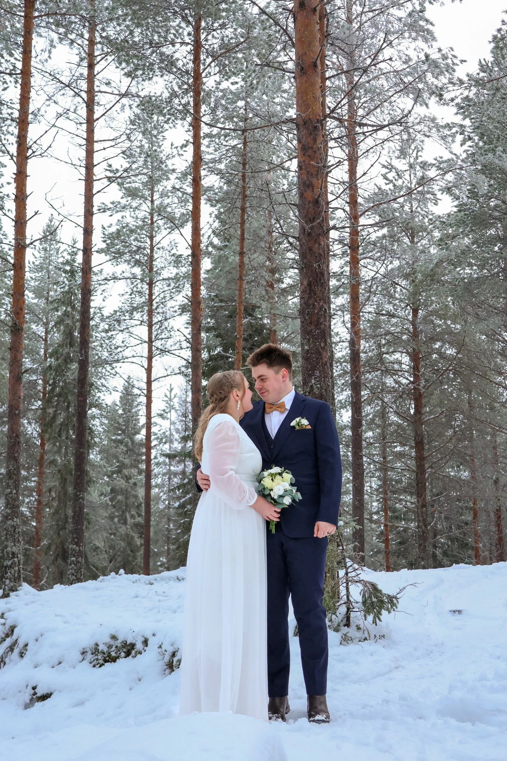 Hääkuvaus hääparista talvisessa metsässä, nainen pukeutuneena valkoiseen hääpukuun ja mies tummansiniseen pukuun keltaisella solmiotupilla. Nainen pitää kukkavihkoa ja molemmat katsovat toisiaan.