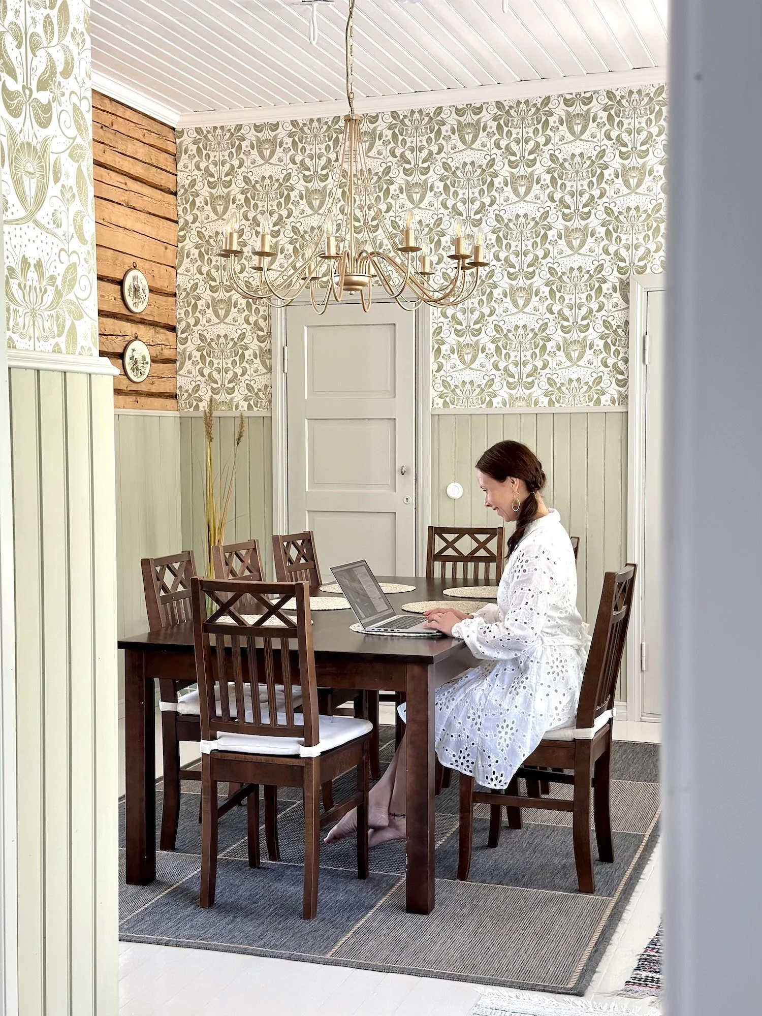 Yrittäjänainen istuu brändikuvauksissaan ruokapöydän ääressä kannettavan tietokoneen kanssa, taustalla on rustiikkinen sisustus ja kattokruunu.