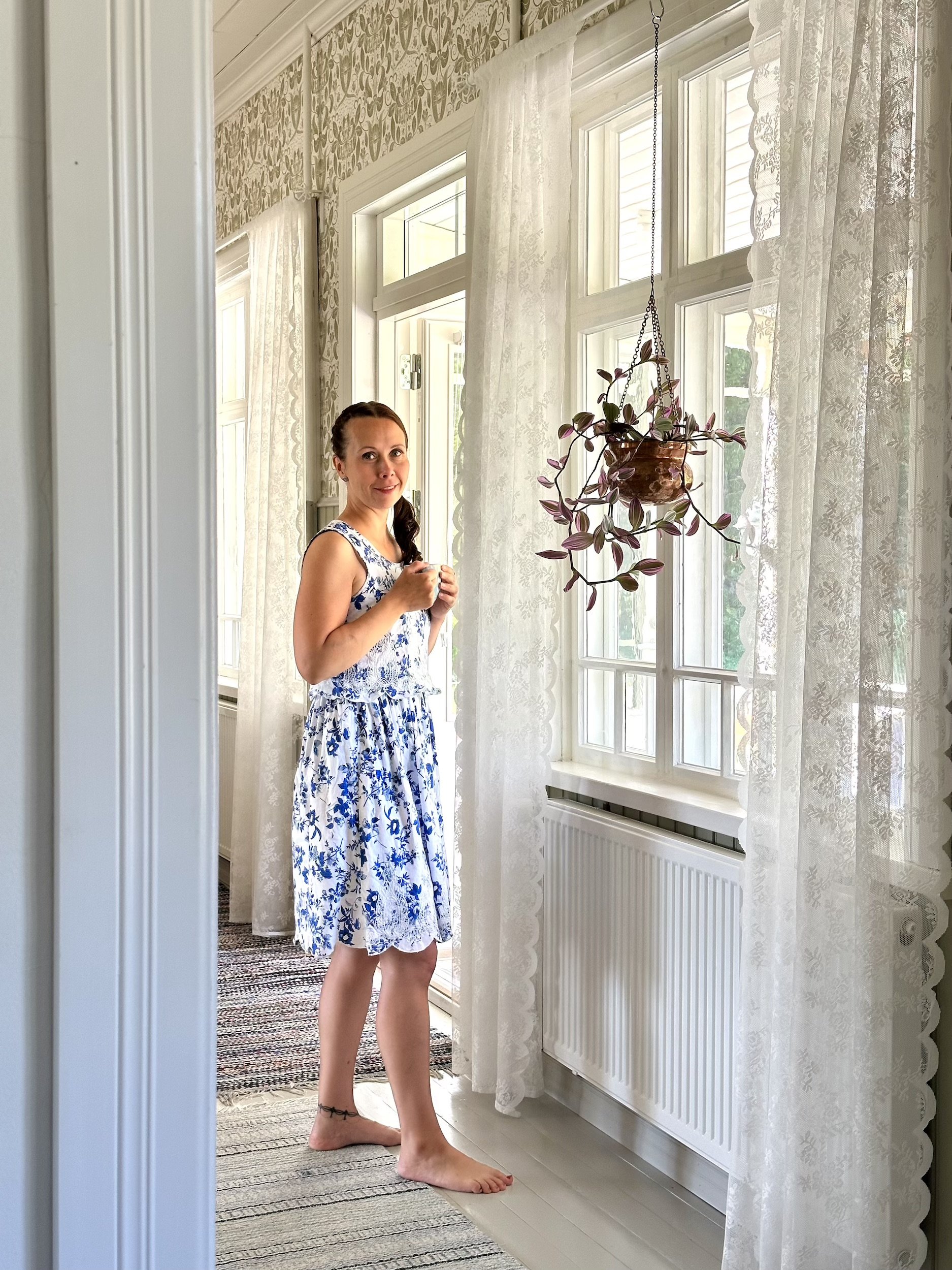 Brändikuvauksessa yrittäjänainen seisoo valkoisessa ja sinisessä kukkamekossa huoneen ikkunan edessä.