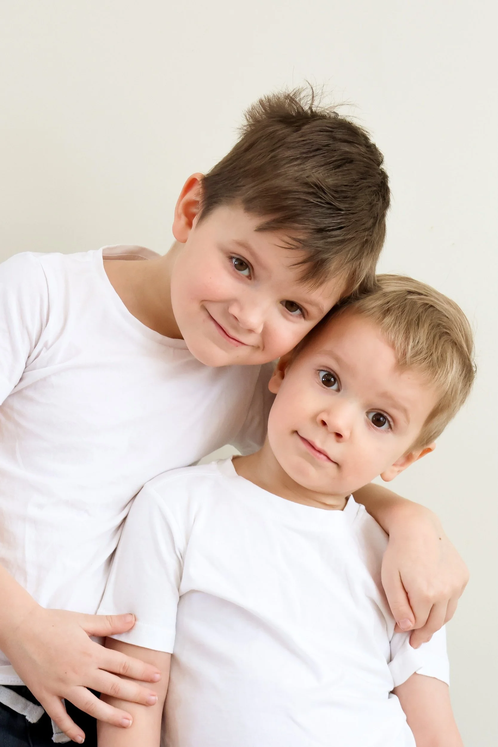 Kaksi lasta hymyilee ja halaa toisiaan valkoisissa t-paidoissa perhekuvauksissa.