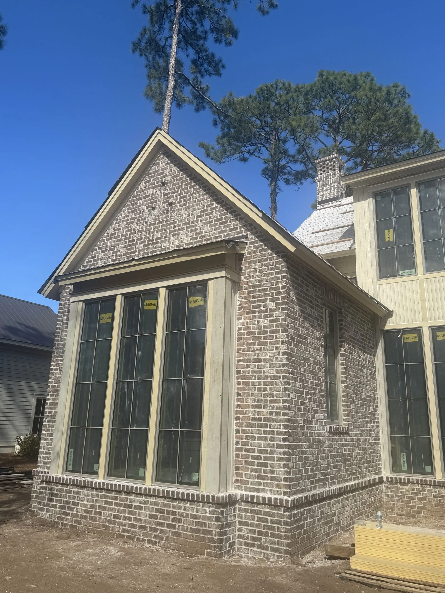 Construction of a brick house with large windows and a gabled roof, surrounded by trees and construction materials on the ground.
