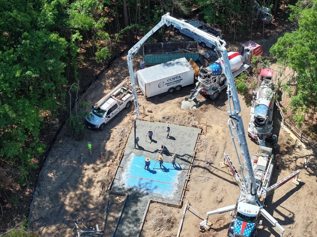A construction site showing workers pouring concrete into a slab with a large cement truck. A concrete pump boom extends over the work area, and there are construction vehicles, including a cement mixer and utility trucks, on the site surrounded by t