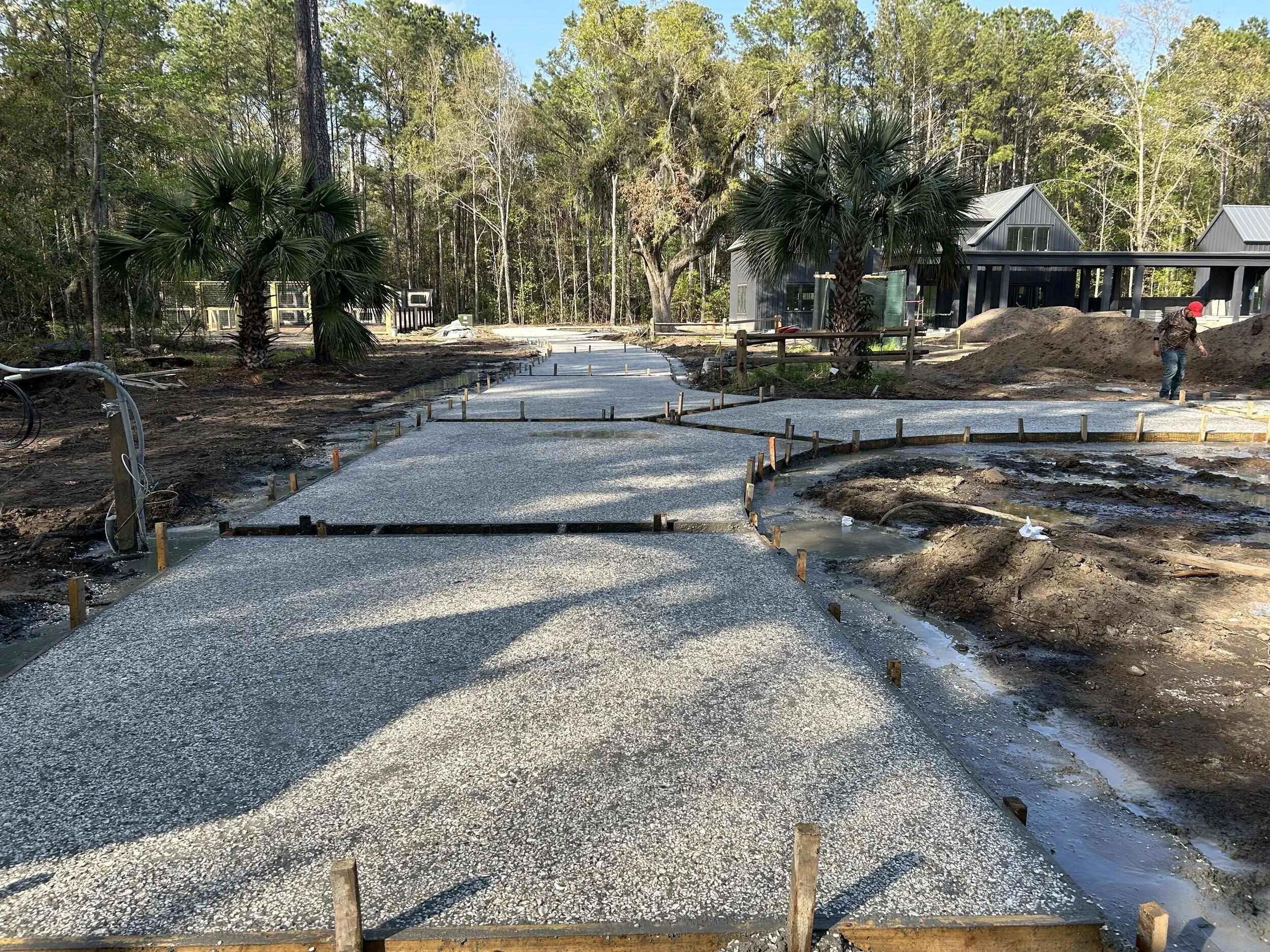 Concrete sidewalk being constructed on a wooded residential property with trees, construction workers, and modern houses in the background.