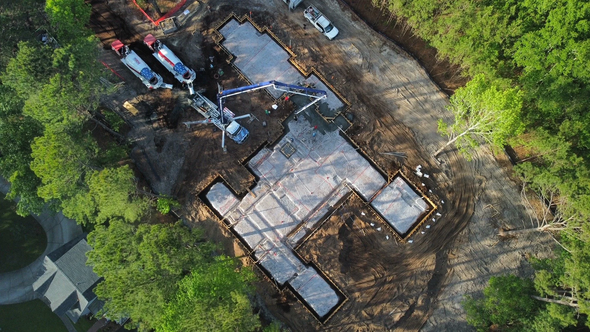 Aerial view of a construction site showing concrete foundation work surrounded by dirt and trees, with construction vehicles on the site.