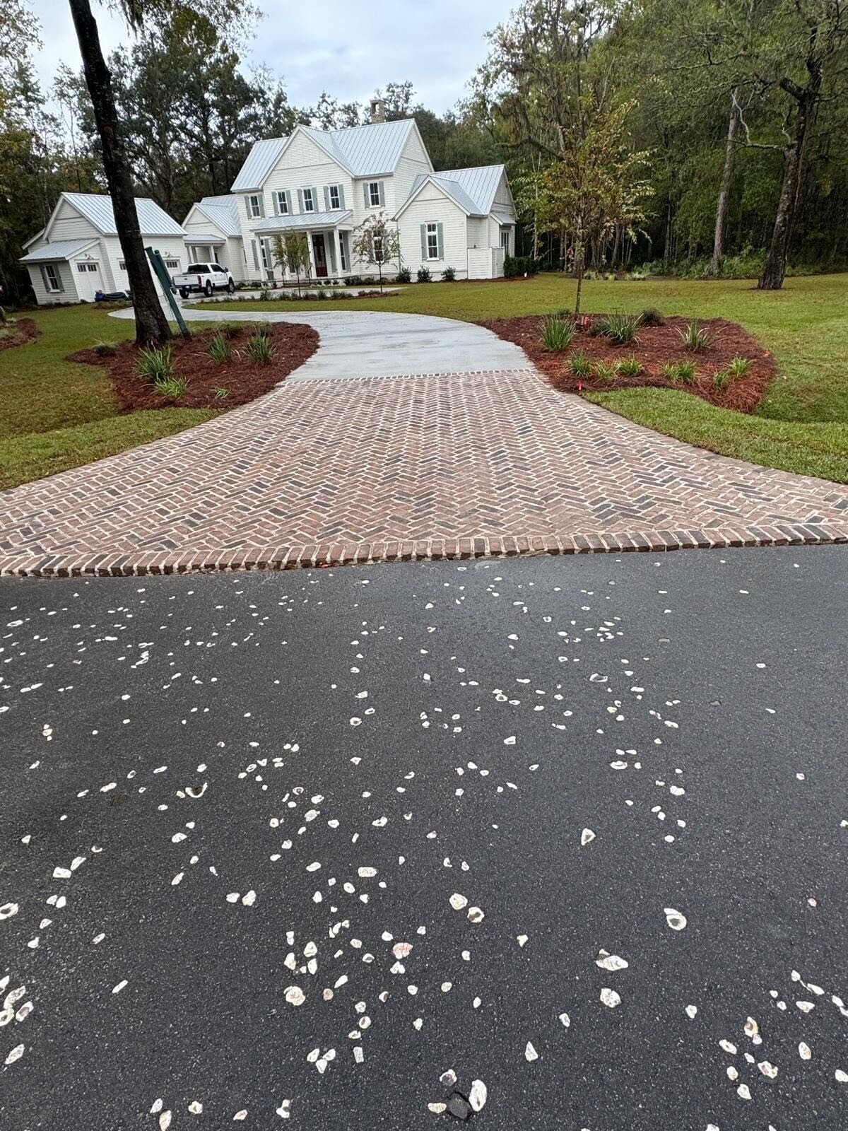 A driveway with brick pavers leading to a white house on a grassy lawn, with landscaped garden beds and trees in the background.