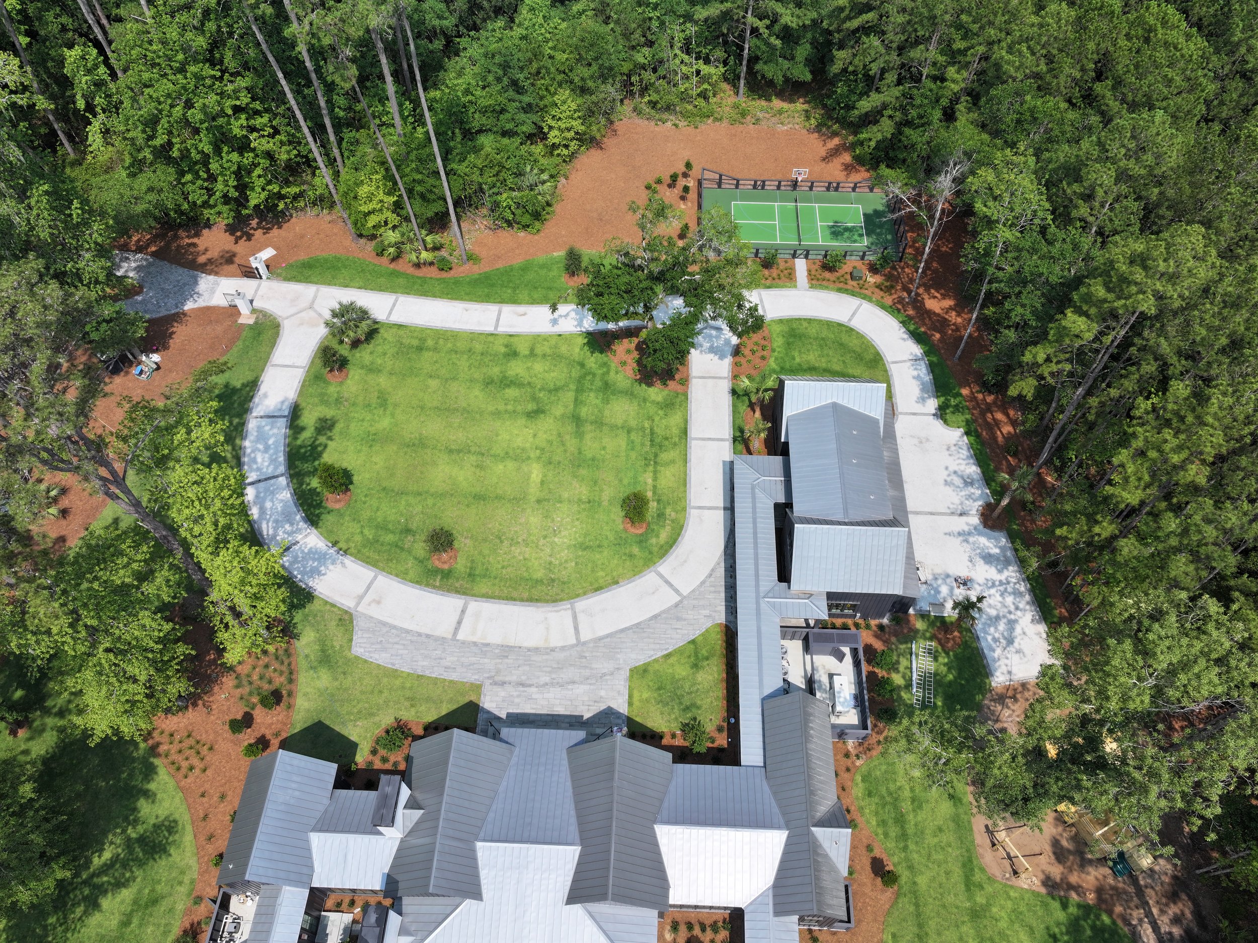 Aerial view of a modern house with a large lawn, connected concrete pathways, a small basketball court, and surrounding trees.