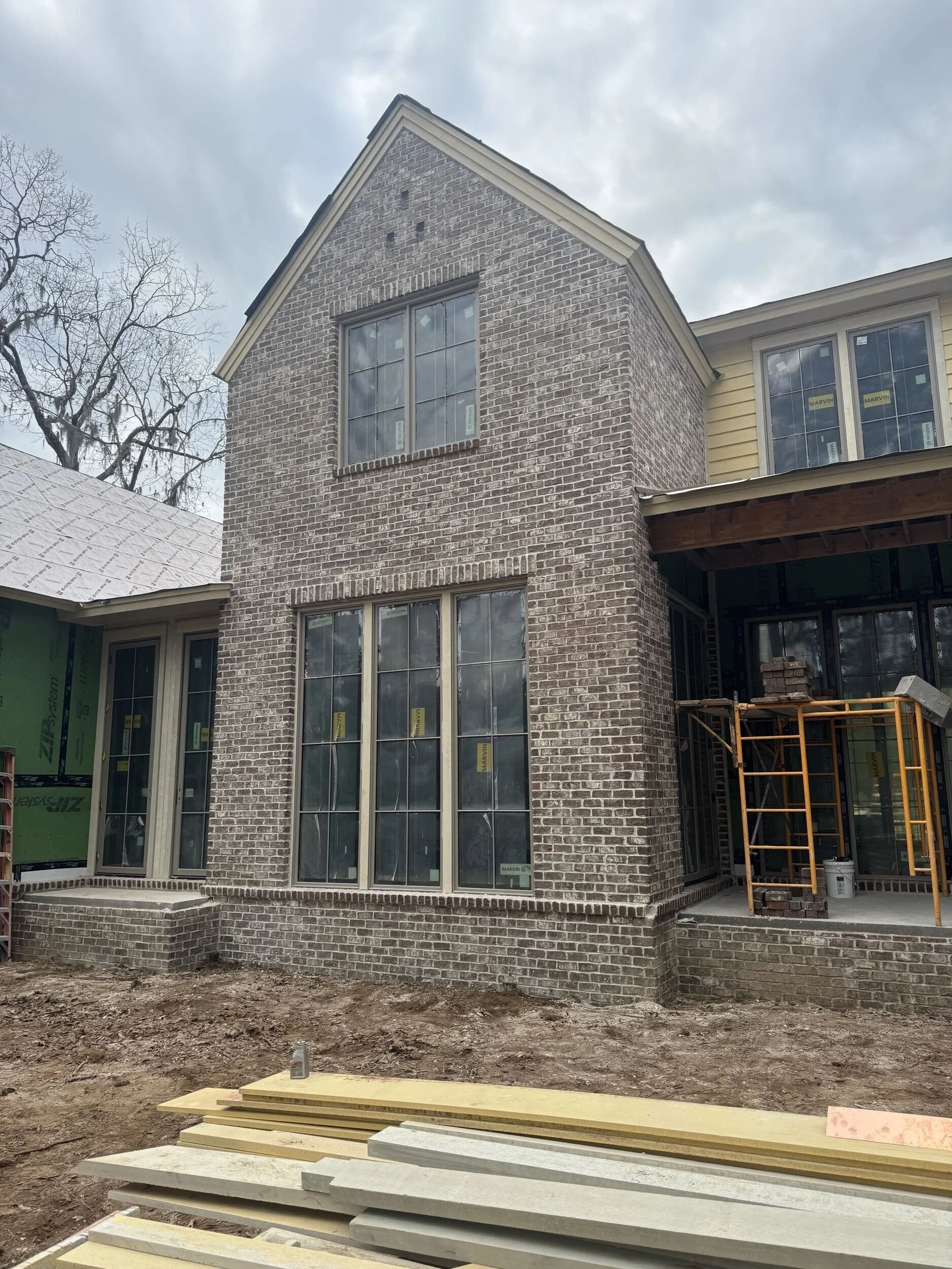 Under construction brick house with large windows and scaffolding, construction materials in the foreground, cloudy sky overhead.