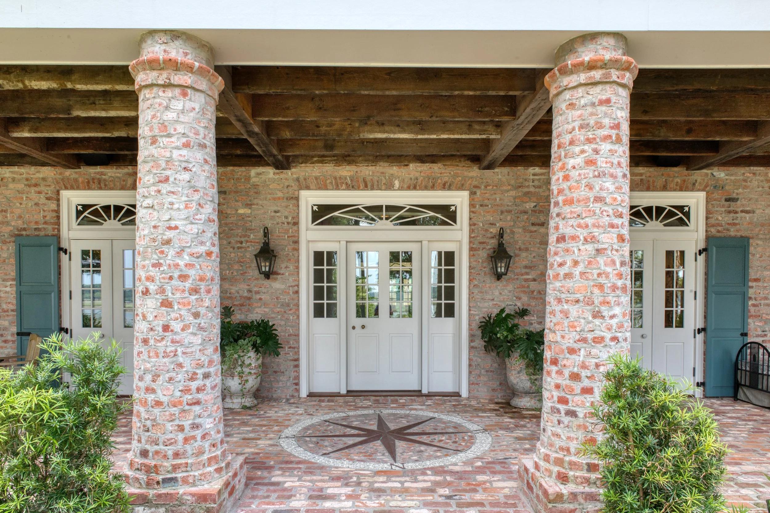 Front porch with brick columns, potted plants, and white double doors with glass panels, flanked by blue shutters and black lantern lights.
