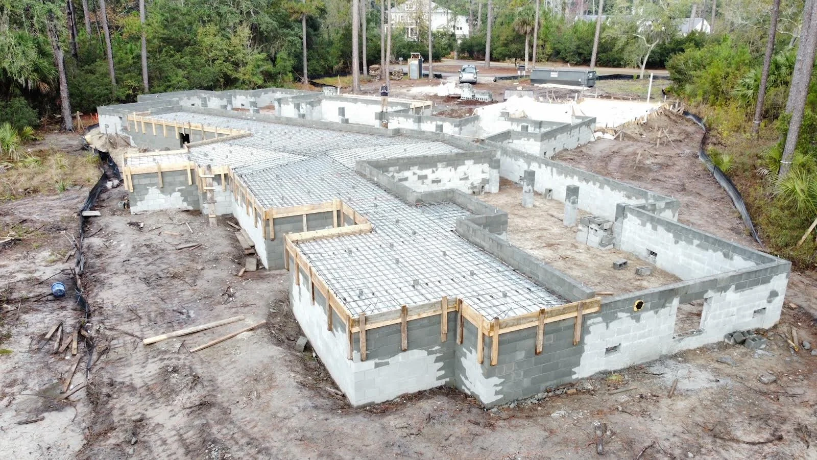 Construction site showing the foundation of a building with concrete blocks, rebar framework, and wooden support structures, surrounded by dirt and trees.