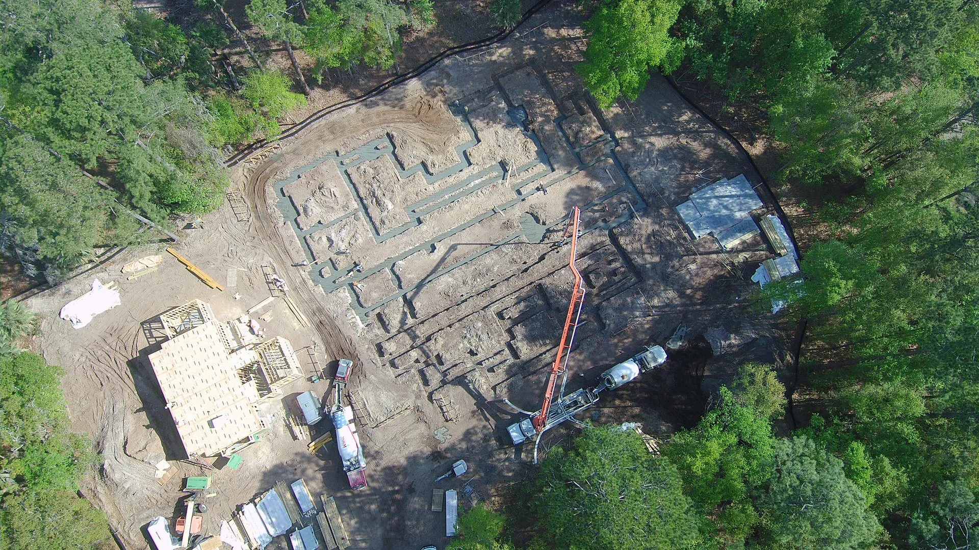 Aerial view of a construction site surrounded by green trees, with early foundations and building framework in progress, a concrete mixer truck, and construction equipment.