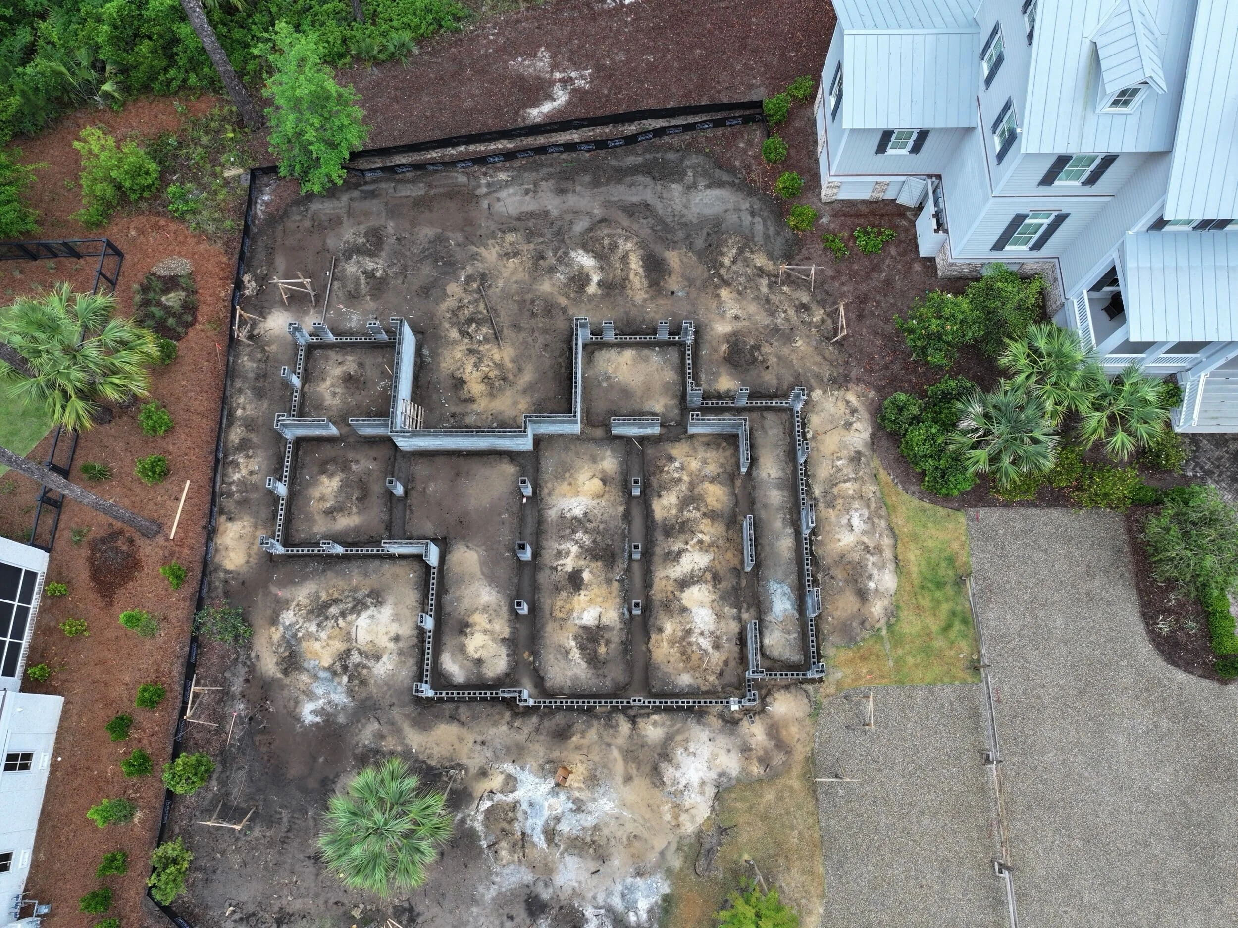 Aerial view of a house under construction with visible concrete foundation framing, surrounded by trees and neighboring houses.