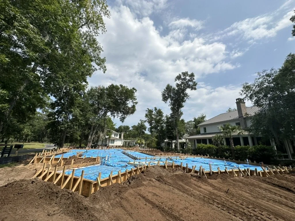 Construction site with wooden framing and blue tarp covering a foundation, surrounded by trees and houses under a partly cloudy sky.