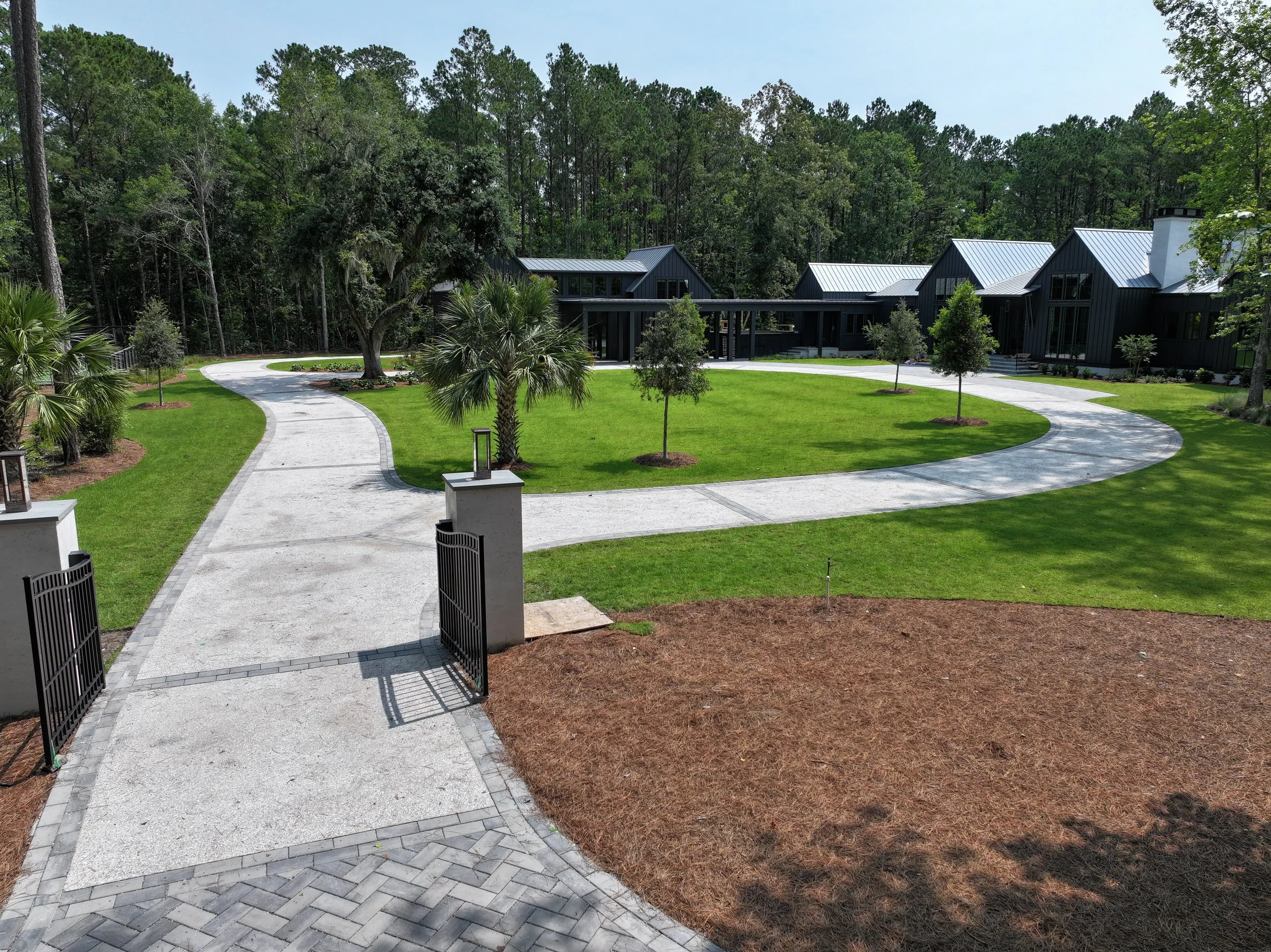 A modern house with dark exterior and metal roof surrounded by a landscaped yard with green grass, small trees, and a curved concrete walking path, set against a background of tall trees.