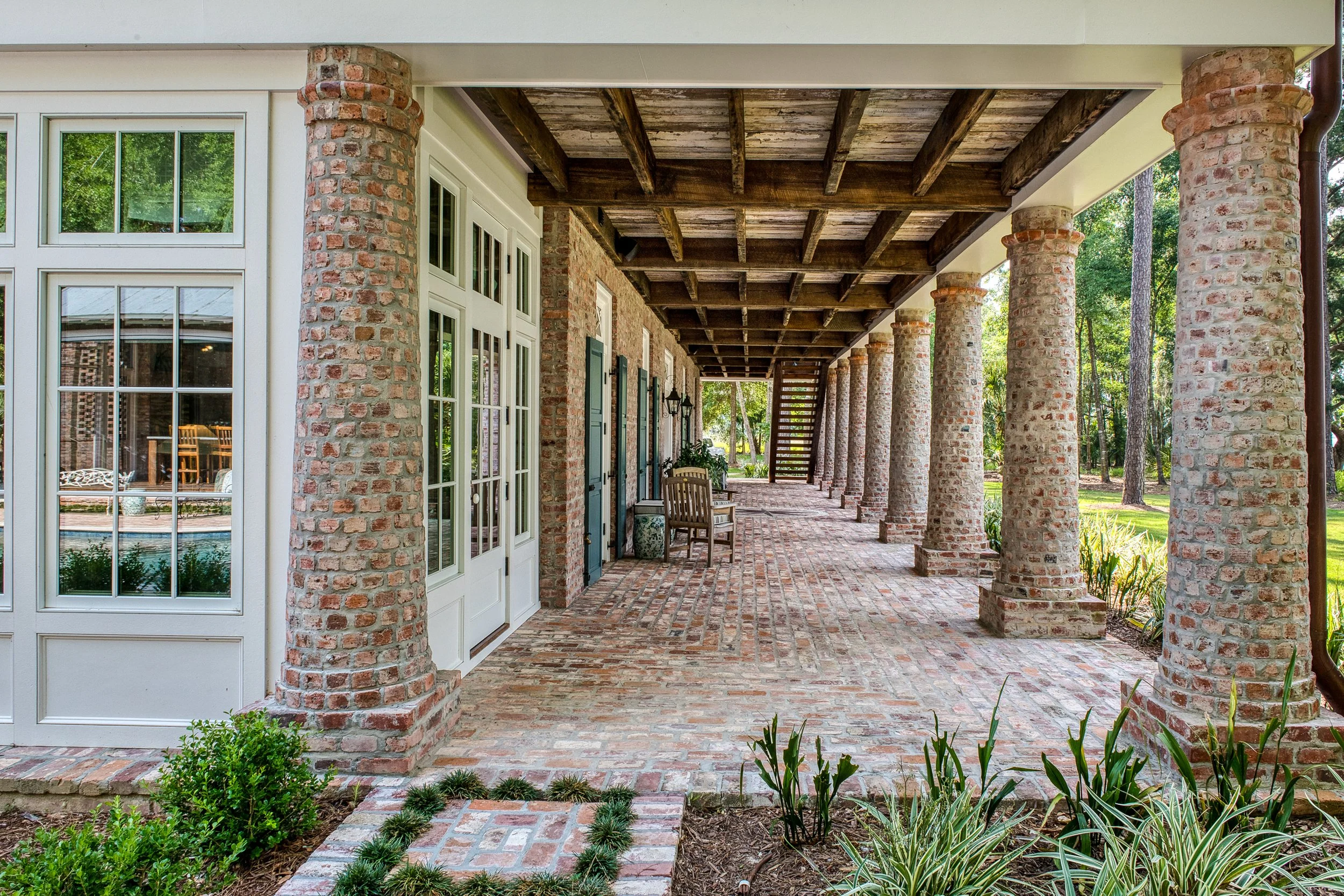 Brick porch with large brick columns, wooden ceiling, benches, and stairs, overlooking a green yard with trees.