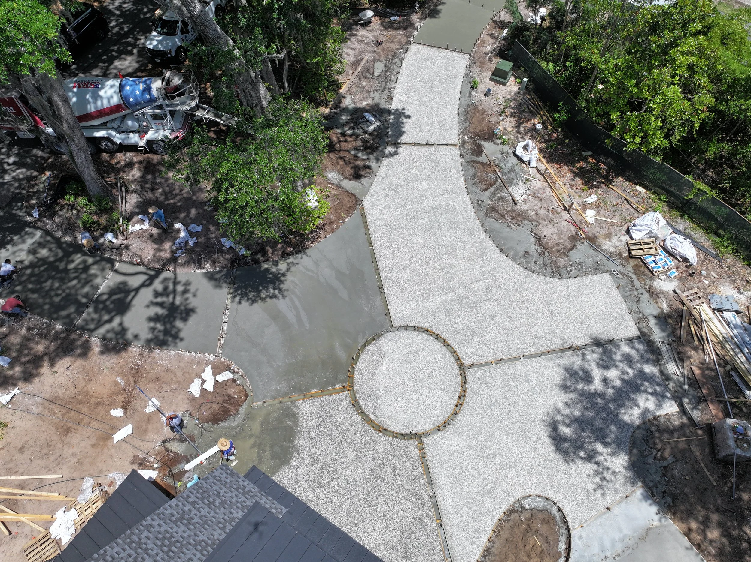 Bird's eye view of a landscaping project with new concrete pathways forming a circular and curving pattern, construction materials on the ground, workers present, and trees providing shade in a backyard.
