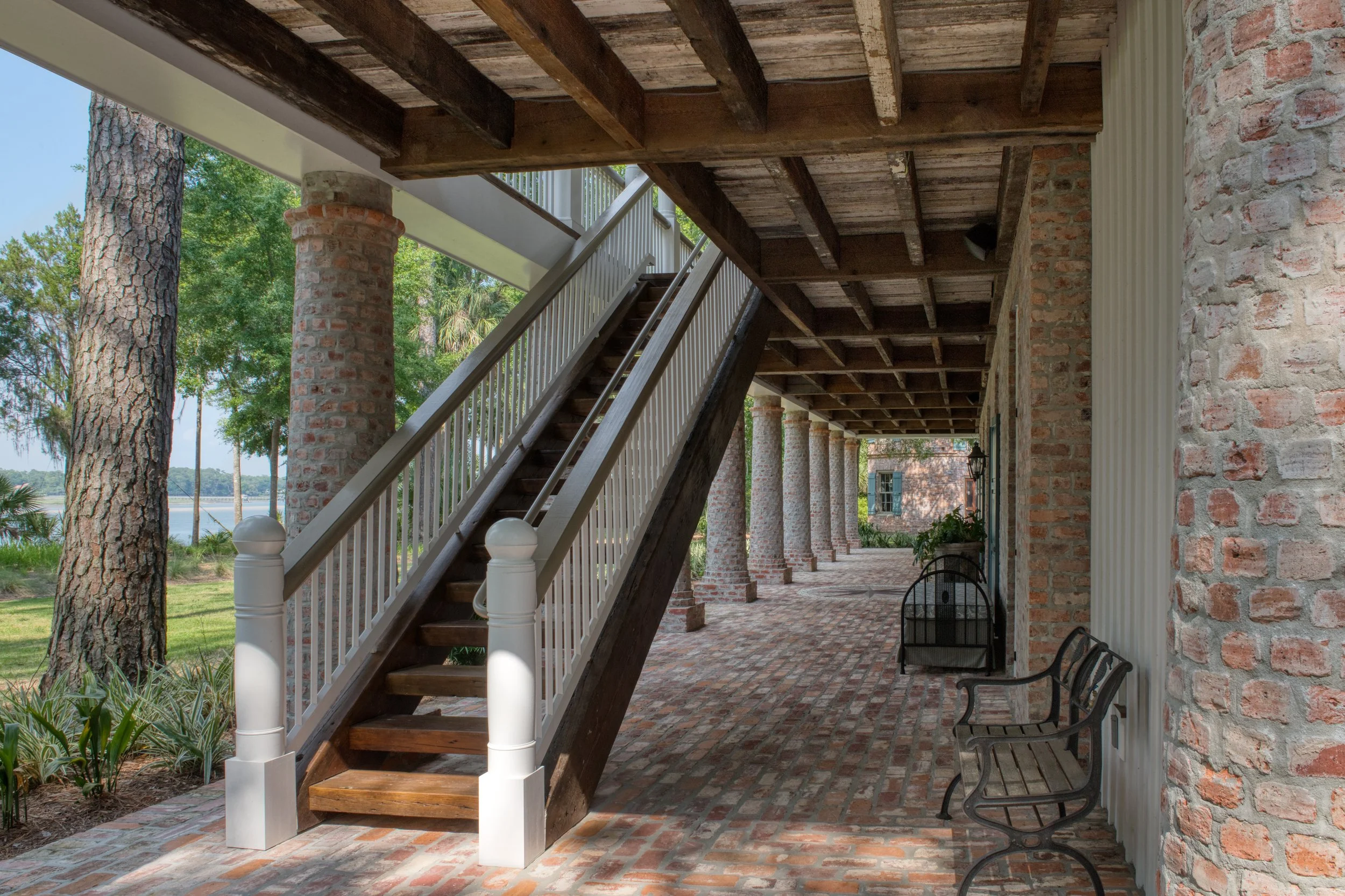 Outdoor porch with brick flooring, wooden ceiling, brick columns, a staircase with white railing, and black metal benches.