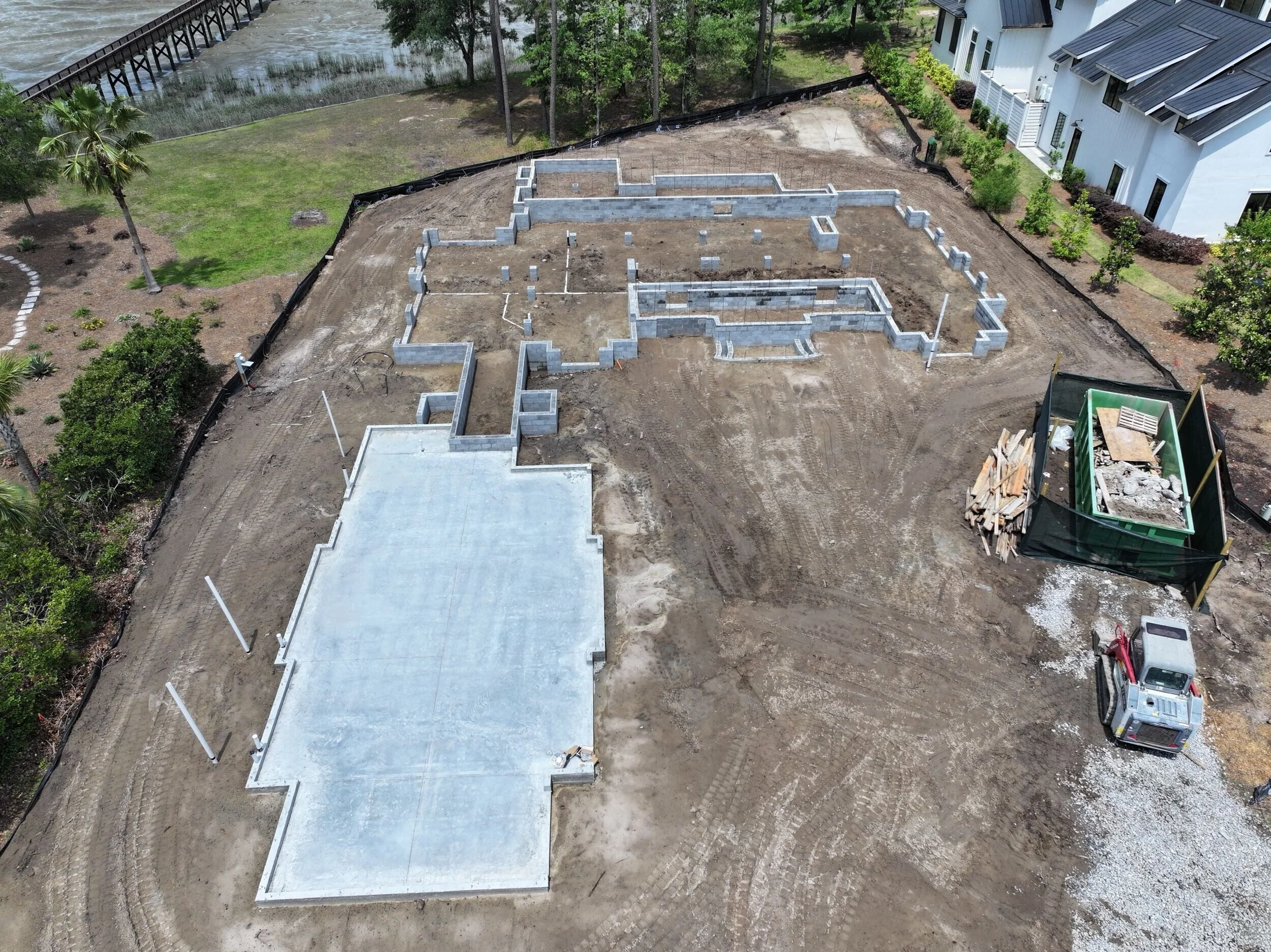 A residential construction site with foundation work in progress, featuring concrete slabs and cinder block walls, surrounded by dirt, construction materials, and a small truck.
