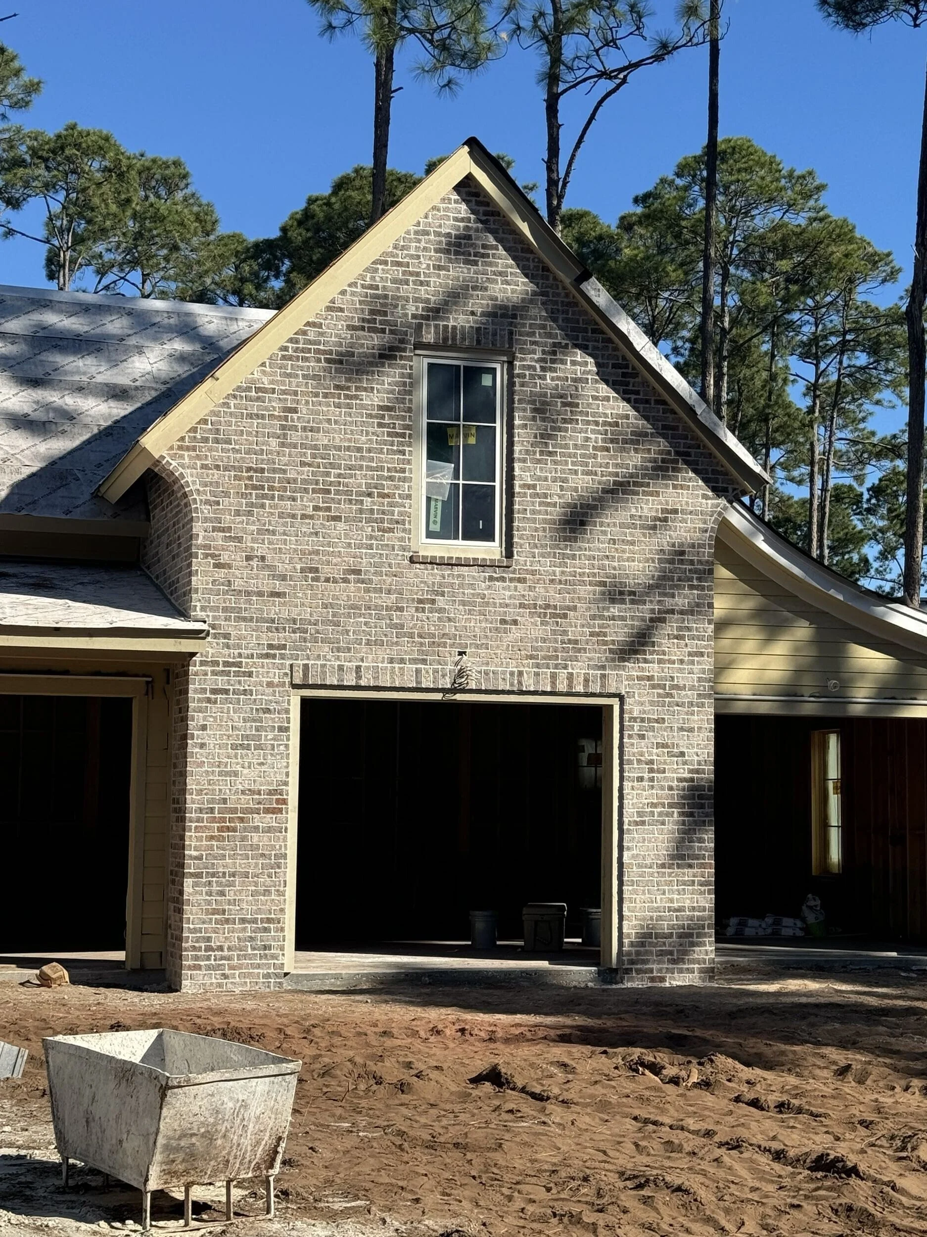 A brick house under construction with an open garage and a yellow side wall, surrounded by tall pine trees and a clear blue sky.