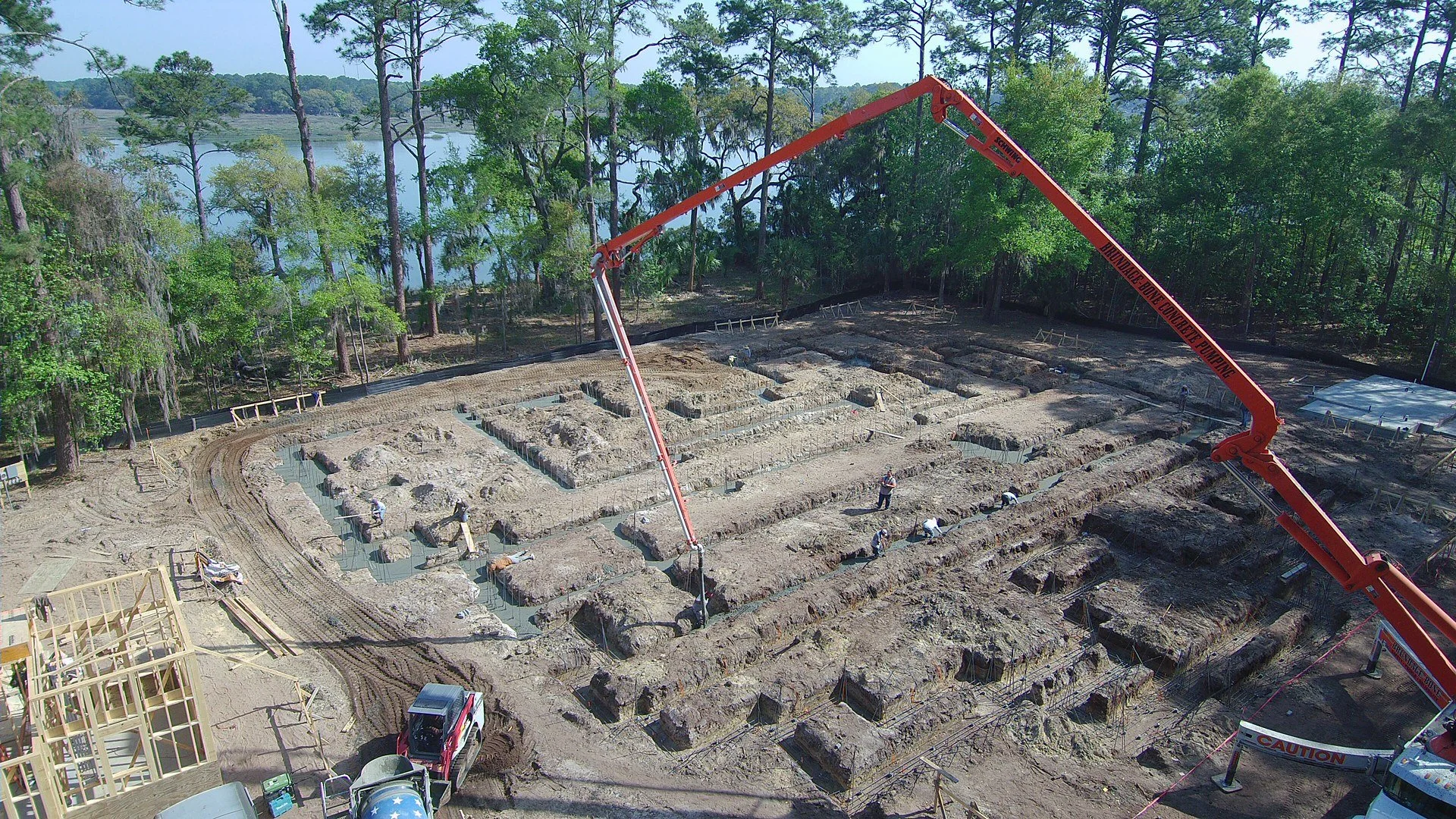Construction site with excavation trenches, workers, concrete pumping truck, and wooden framing structures surrounded by trees and a lake in the background.