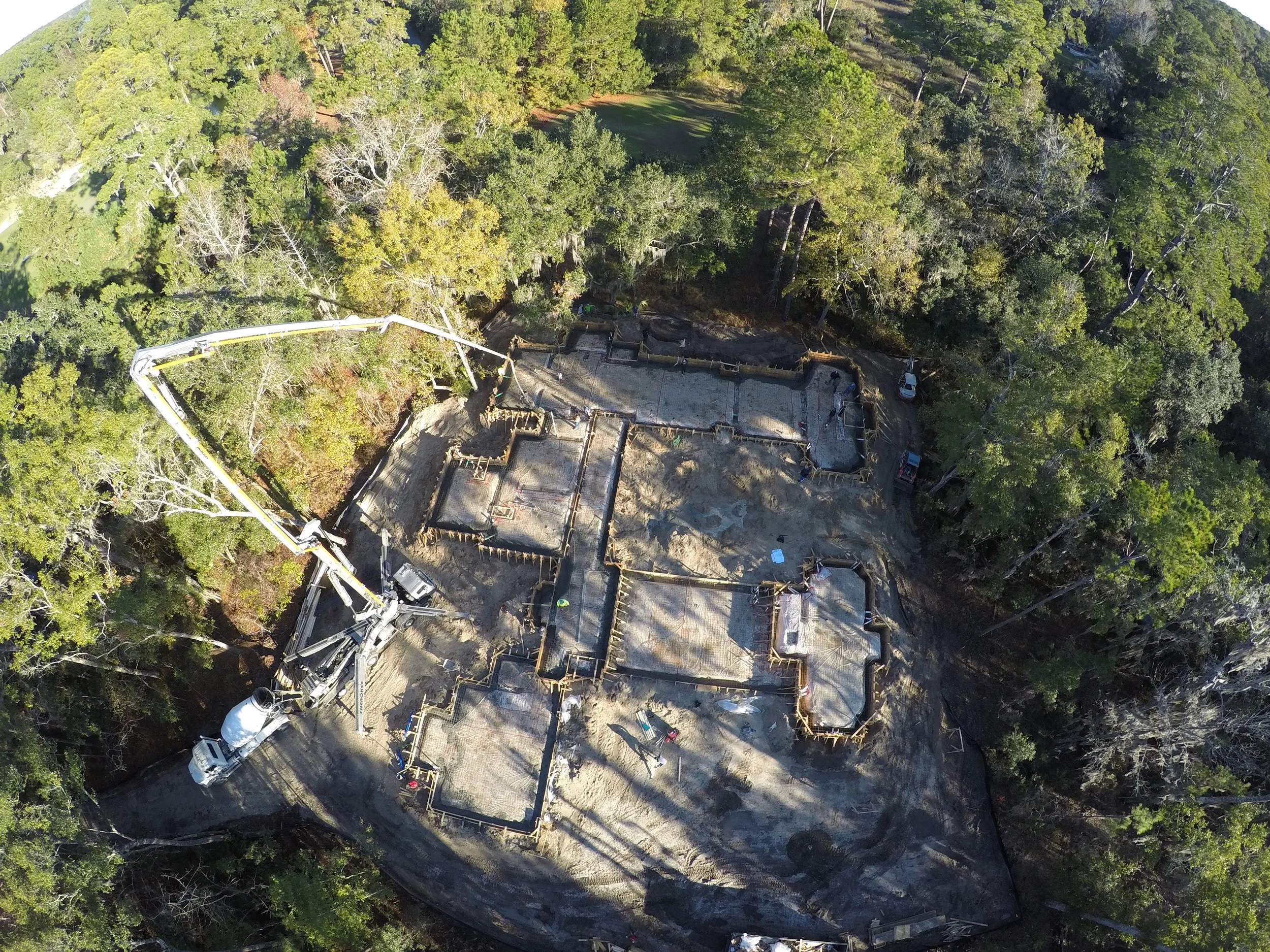 Aerial view of a construction site in a wooded area with building foundation work and concrete pouring in progress, surrounded by green trees.