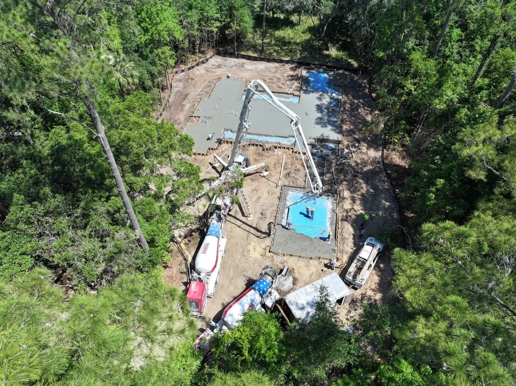 A construction site in a wooded area shows concrete being poured for a building foundation, with construction workers, a cement mixer truck, and a concrete pump truck visible.