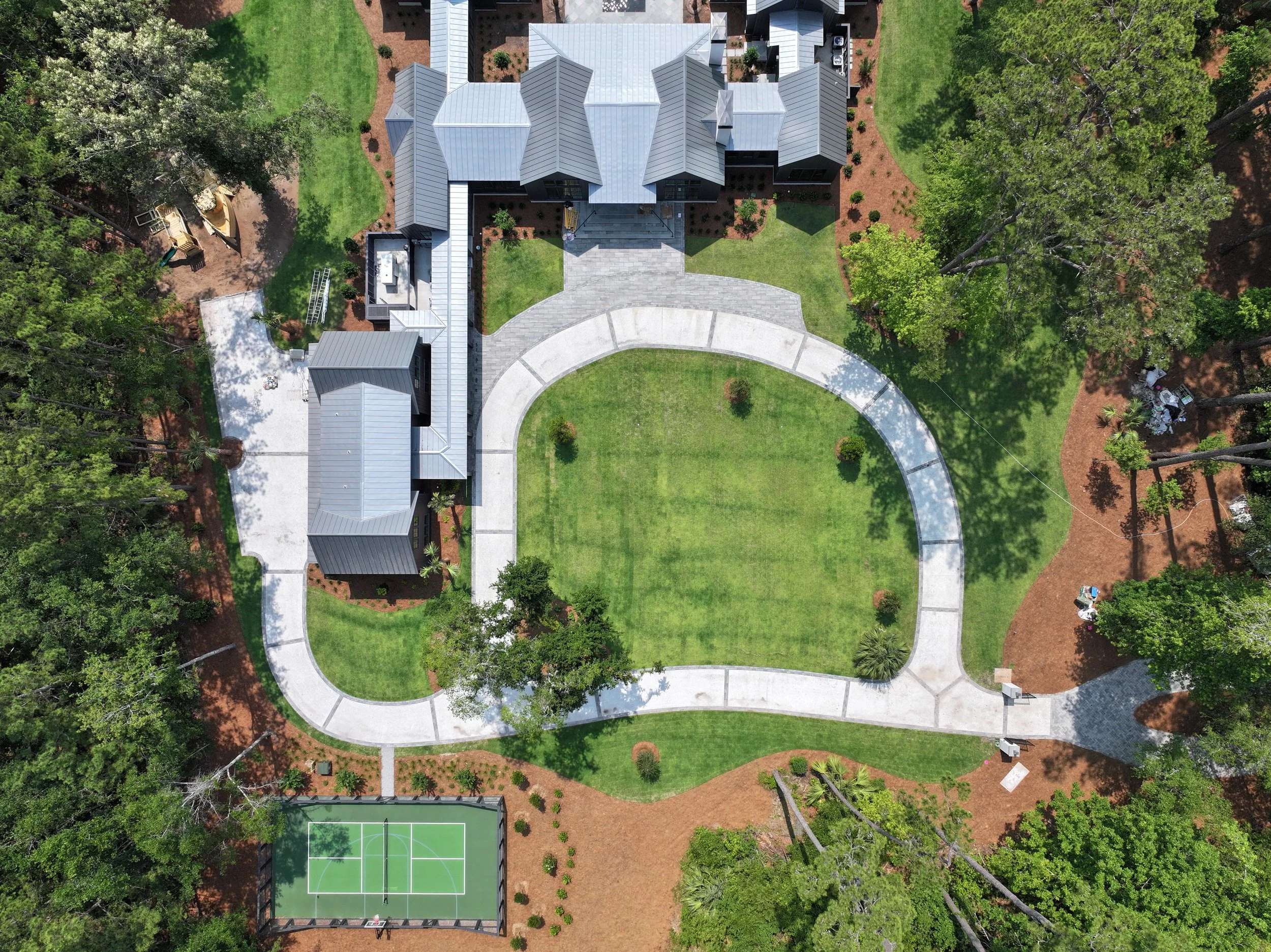 An aerial view of a large house with a gray roof, surrounded by a well-maintained lawn, trees, and pathways. There is a tennis court at the bottom of the image and a playground to the left. The landscape is lush and green.
