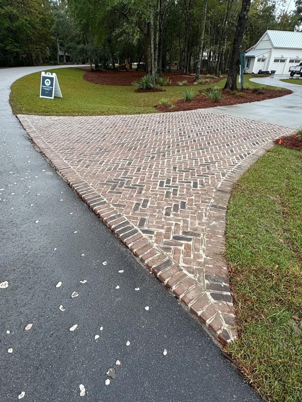 A driveway and sidewalk intersection with brick paving, grass, and trees in a residential neighborhood.