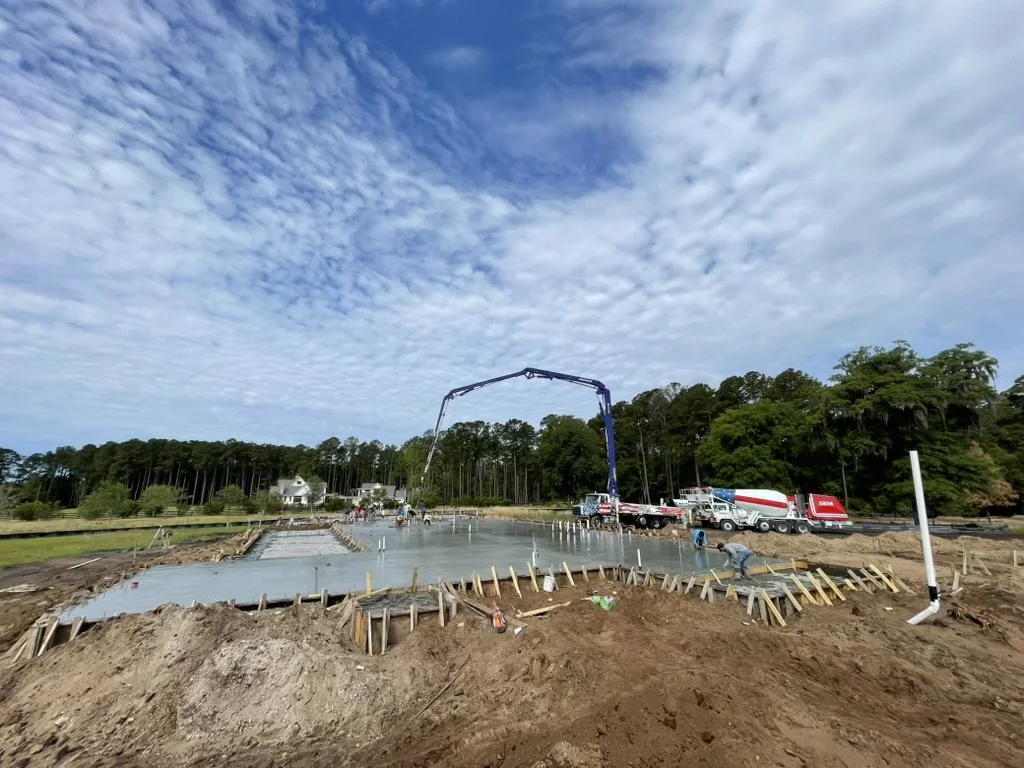 Construction site with concrete being poured for a building foundation, surrounded by trees and a blue sky with clouds.