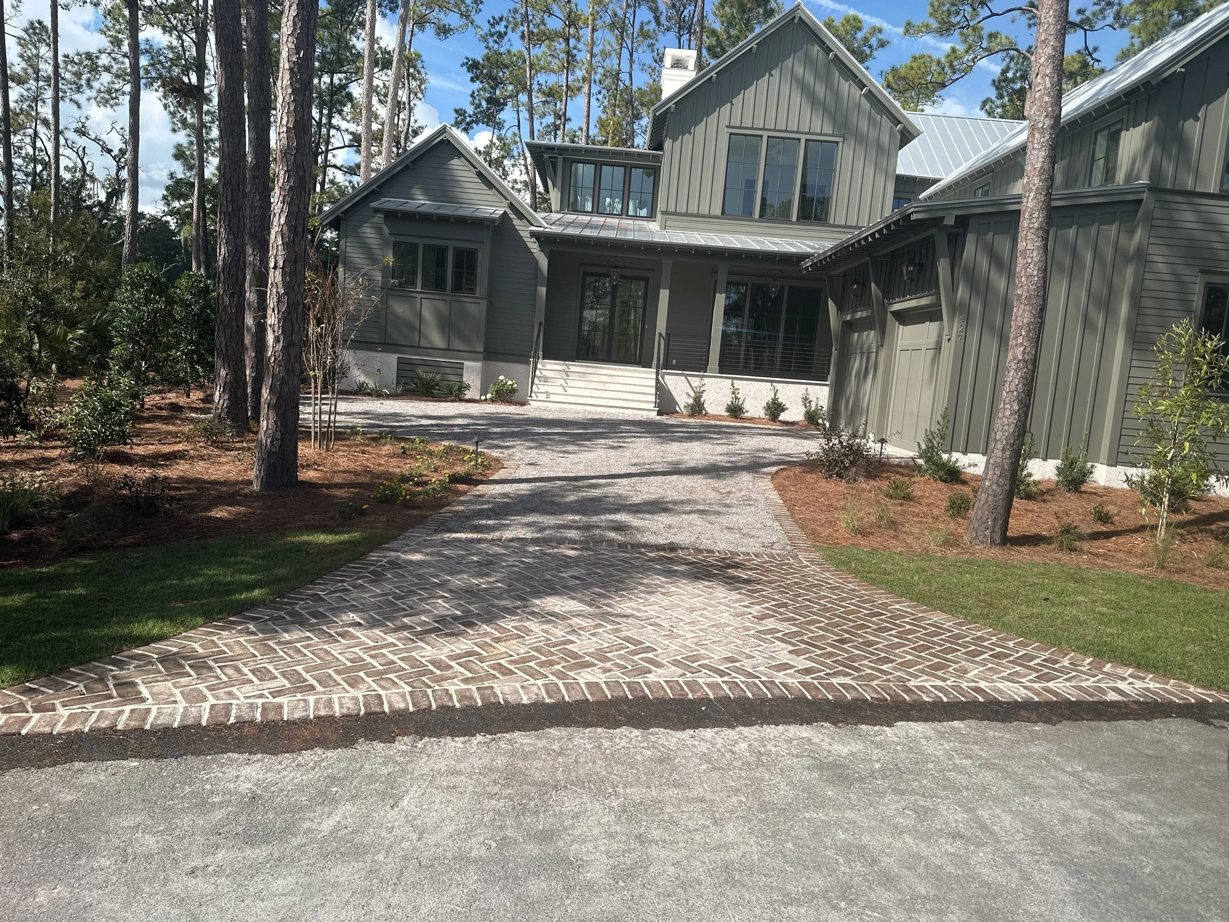 Modern house with a gray exterior, large windows, and a sloped metal roof, surrounded by trees and landscaped yard with a brick and gravel driveway.