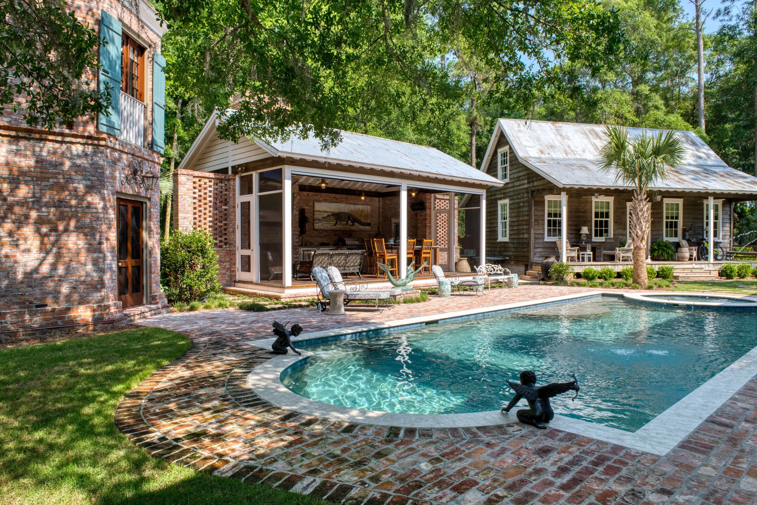 A backyard swimming pool surrounded by brick and stone paving, with decorative frog statues at the edge, adjacent to a covered outdoor sitting area and two different styled houses, one with brick exterior and the other with wooden siding, amid green 