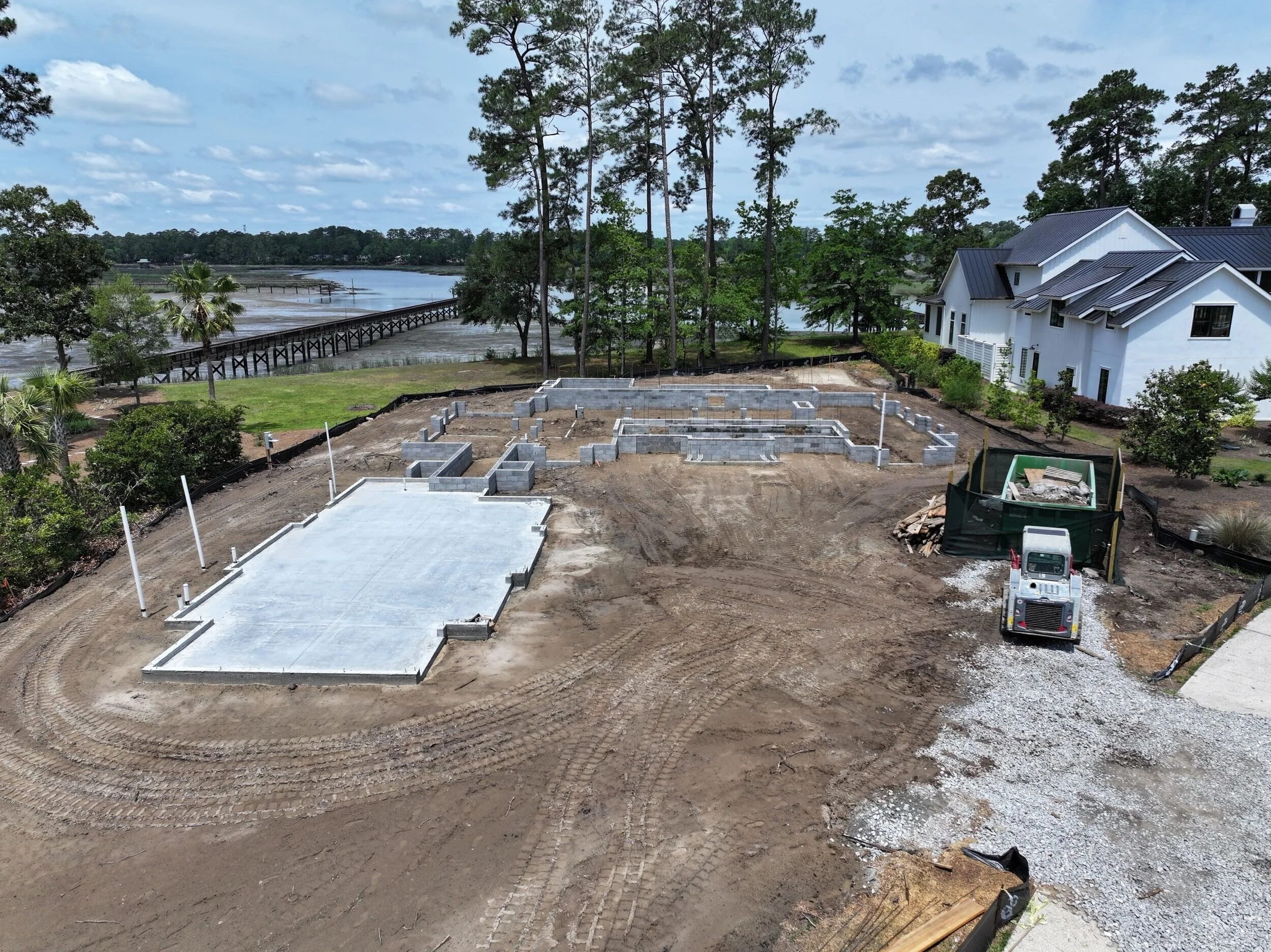 Construction site with foundation and early building framing near a river with trees and neighboring white house.