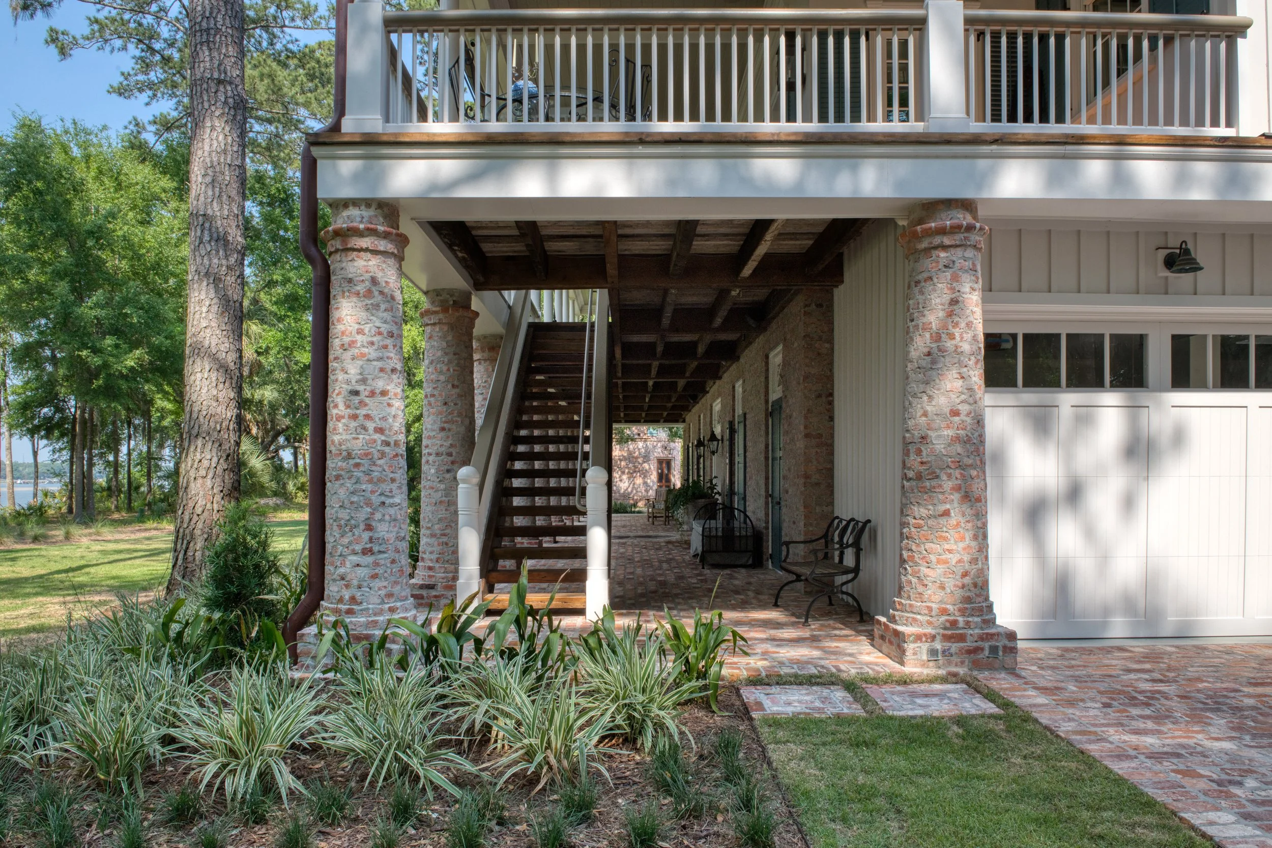 The back porch of a house with brick columns, a staircase leading to a second-floor balcony, outdoor seating, and a well-maintained garden with trees and grass.
