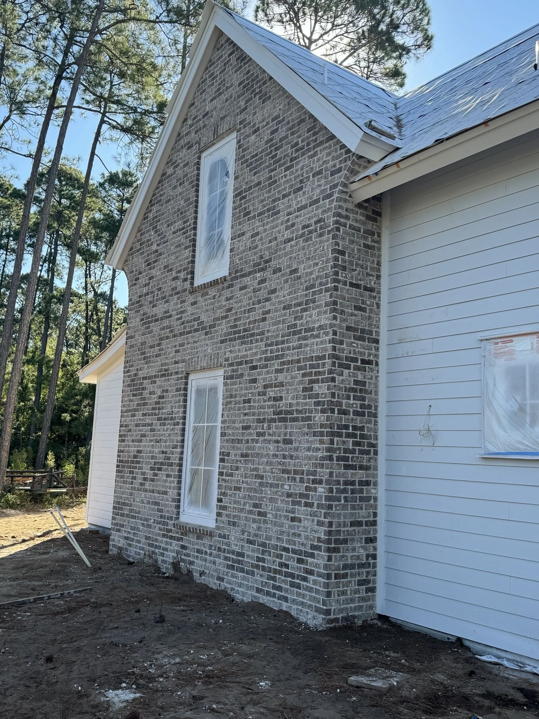 Side view of a house under construction with a wood and brick exterior, wrapped windows, nearby trees, and unfinished ground.