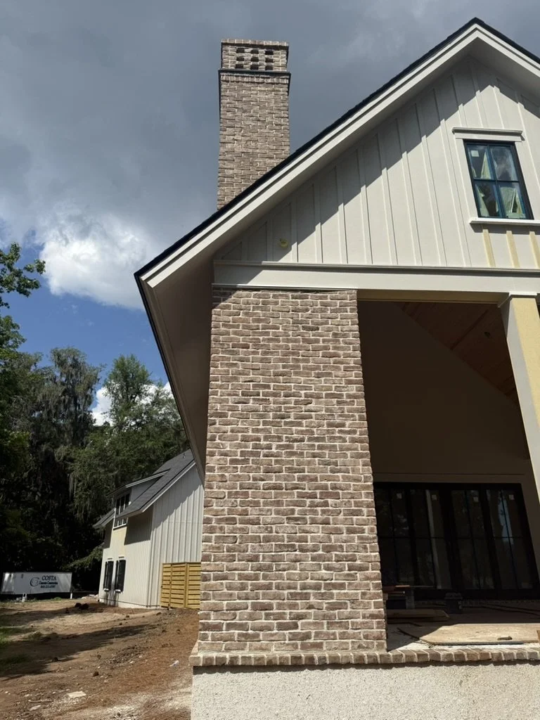 Close-up view of a brick chimney attached to a modern house with white exterior siding and large glass doors, under a partly cloudy sky.