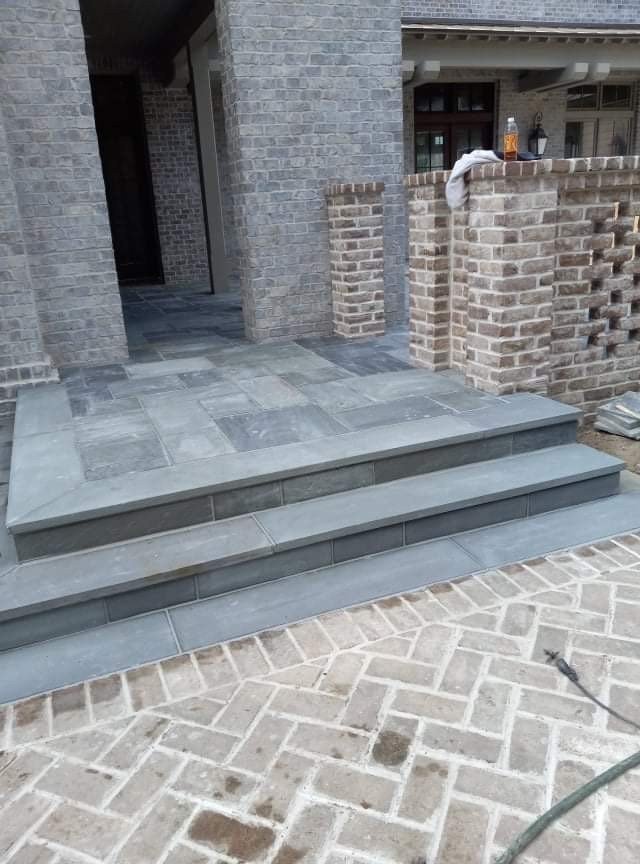 Exterior view of a building entrance with a few steps leading up to a porch. The steps are made of gray tiles, and the porch has a gray tiled floor. The building is made of gray and brown bricks, with a wrapped white cloth and a bottle placed on the 