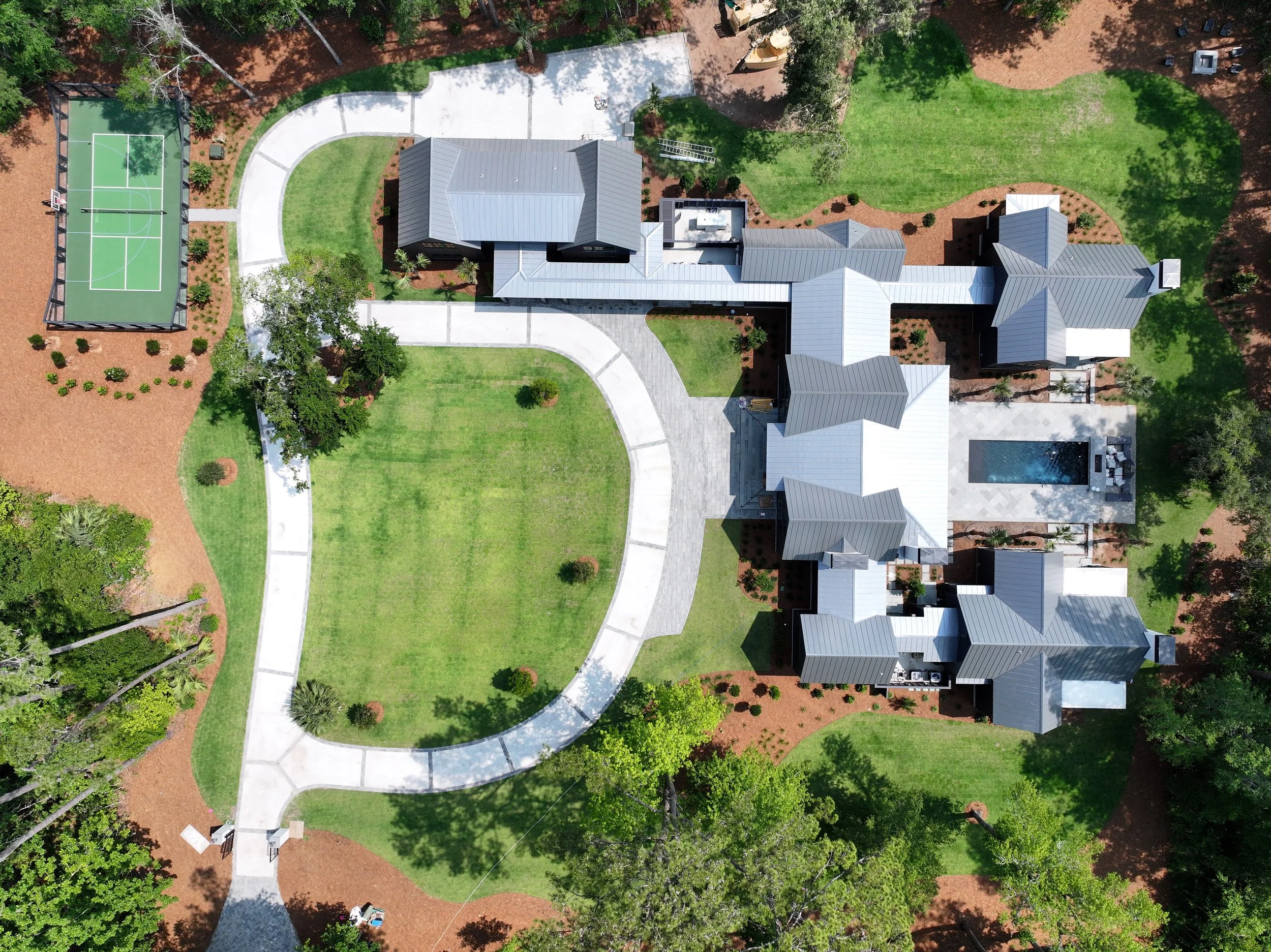 Aerial view of a large modern house with gray metal roof and a backyard featuring a swimming pool. The house is surrounded by green lawns, trees, and landscaped gardens. There are paved walkways around the property, a tennis court on the upper left, 