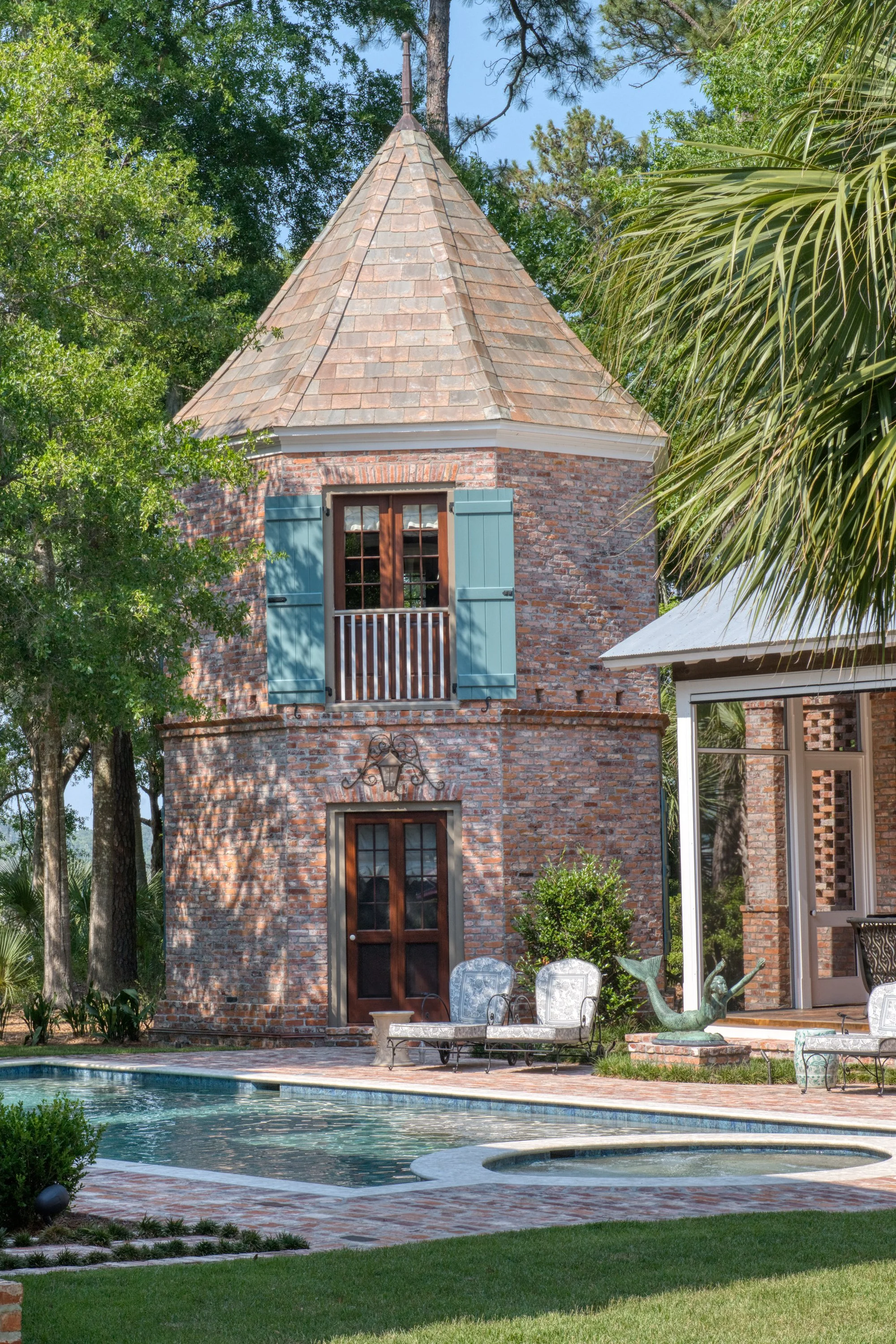 A brick house with a round tower featuring a shingled conical roof, blue wooden shutters, a small balcony, and a door beneath a decorative wrought iron piece. In front, there is a swimming pool with chairs, a fountain, and lush green trees surroundin