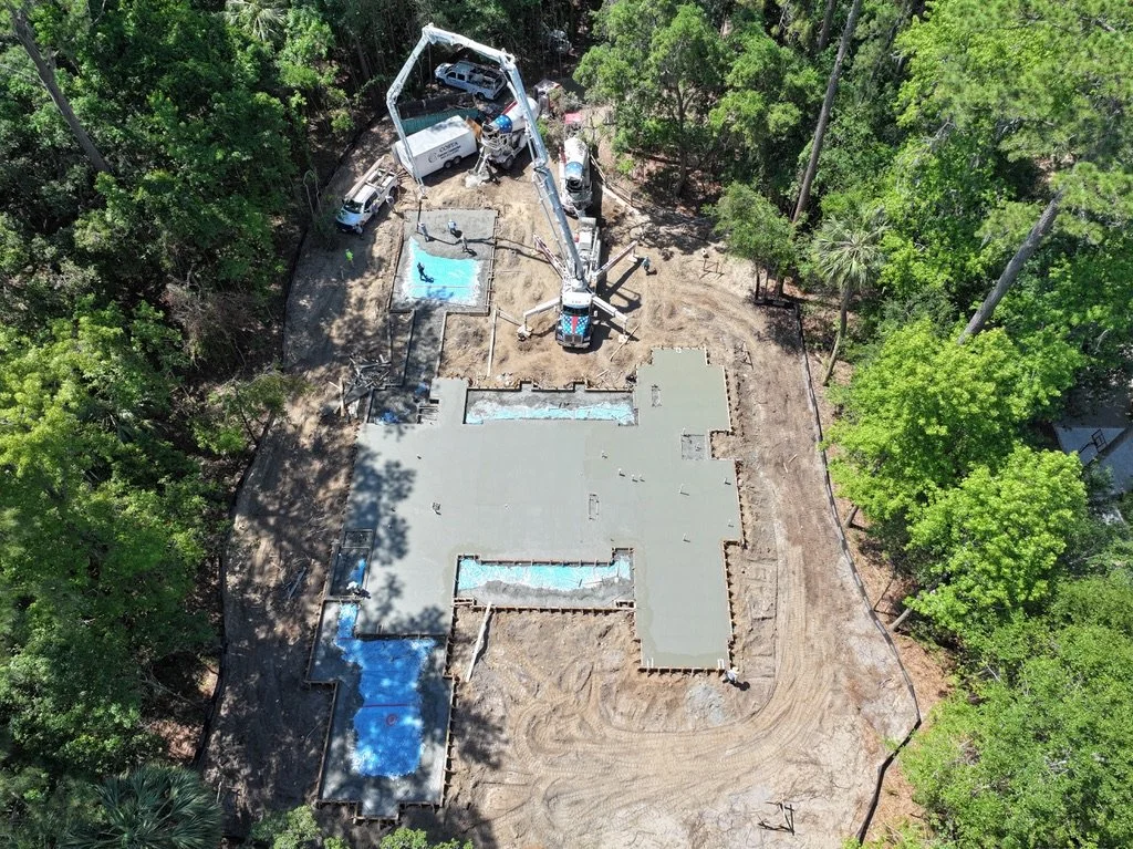 An aerial view of a construction site where a house is being built, with concrete being poured over the foundation, surrounded by green trees and construction equipment.